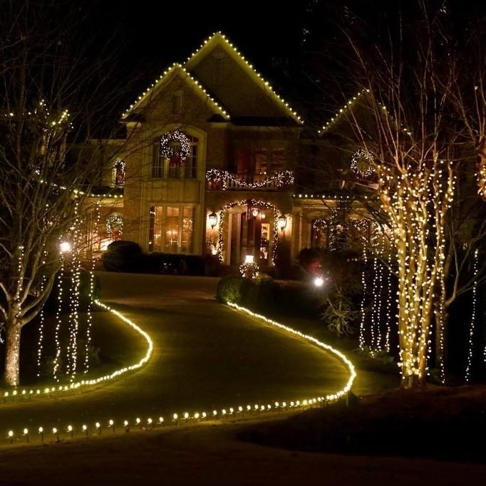Home at night behind an S-curve driveway edged in warm white, with mature trees on both sides wrapped from trunk to branch tips