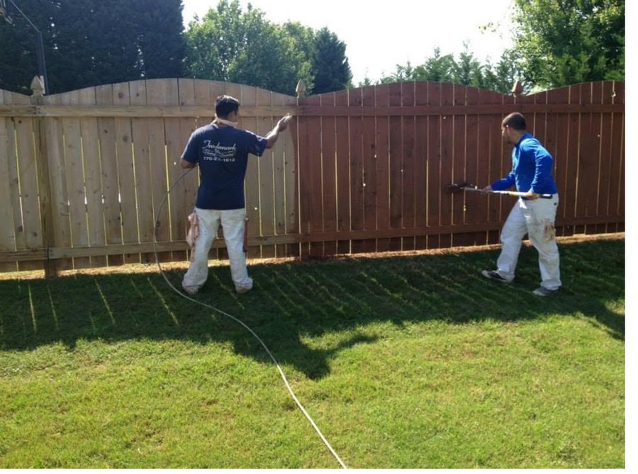 Two Trademark Design crew members in branded shirts spraying and back-brushing stain onto a timber fence